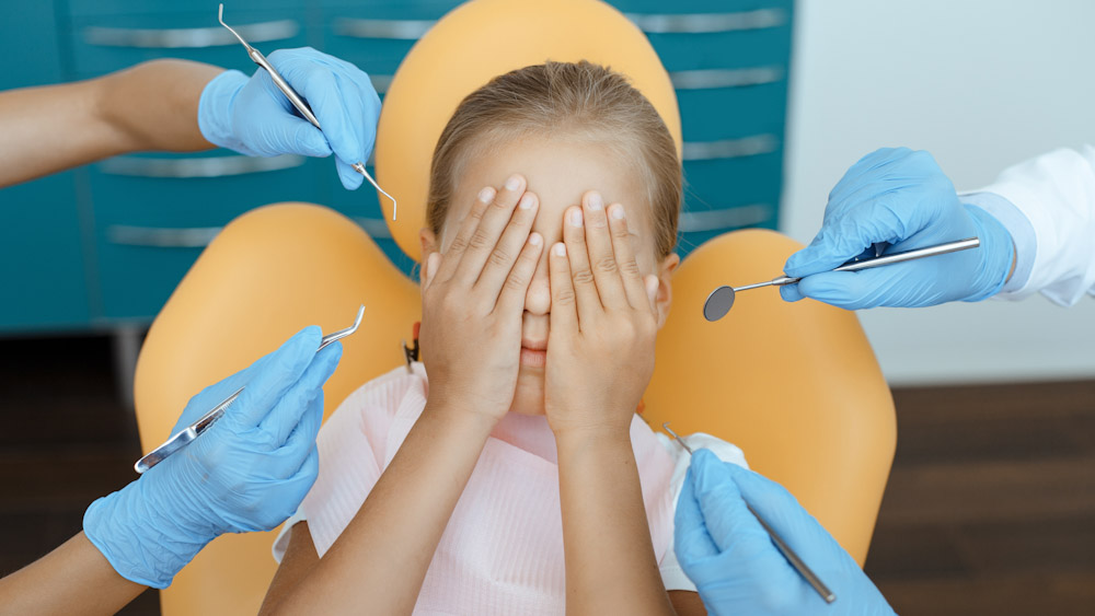 A photo of a child at the dentist, covering her eyes with nervousness as gloved hands approach.